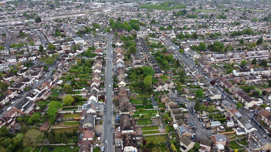 An aerial view of a residential area within the Alton Estate in Roehampton, showing a mixture of low-rise terraced houses and high-rise apartment buildings. The scene captures a street with parked car